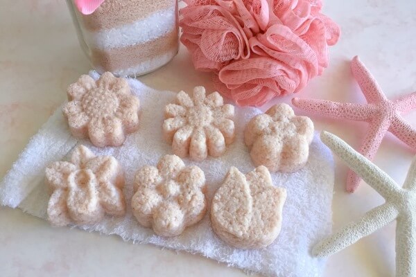 pink himalayan and epsom salt cakes in the shape of flowers laying on a face washer surrounded by starfish and a pink loofah sponge