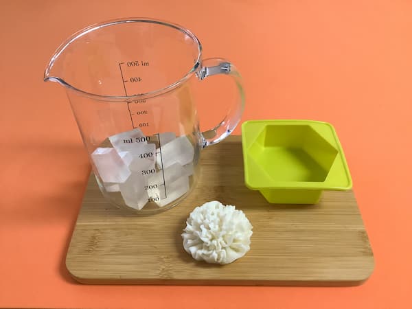 a white carnation-shaped soap bar sitting next to a glass jug filled with clear soap base and an empty silicone mould in the shape of a hexagon