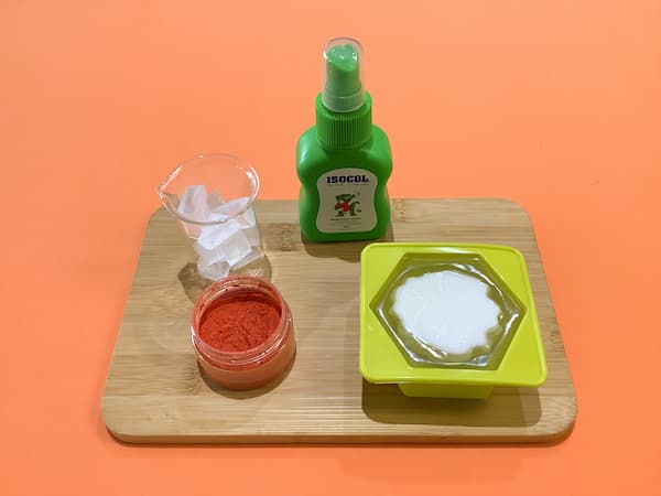 a glass beaker filled with clear soap base sitting next to a bottle of rubbing alcohol, a container of orange mica, and a hexagonal soap mould filled with clear soap base and an upside down carnation soap bar