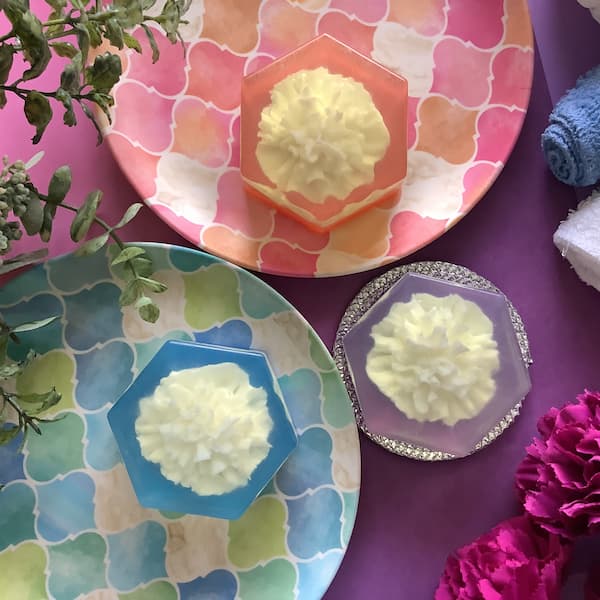 three embedded carnation melt and pour soap bars, each with a white carnation against a blue, purple or orange hexagonal background